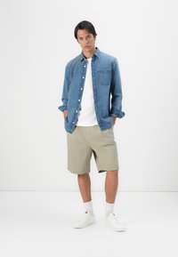 Denim overhemd, lichtblauw, met knopen en een borstzak. Witte t-shirt eronder, gecombineerd met beige shorts. Witte sneakers en sokken.