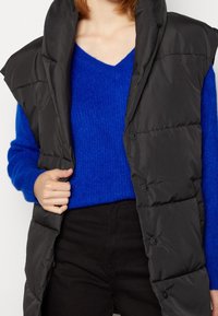 Veste matelassée noire avec un col montant et une fermeture à boutons-pression, portée par-dessus un pull bleu en tricot. Présente un design élégant et une texture douce.
