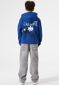 Sudadera azul con un estampado gráfico que presenta flores y formas abstractas. Combinada con pantalones vaqueros de corte holgado en gris claro. Material de algodón, diseño casual.