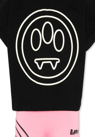 Chemise noire avec un visage abstrait en contour blanc, présentant trois yeux ovales et deux petites crocs, associée à un pantalon rose avec du texte et des lignes noires.