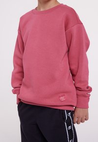 Bambino in felpa rosa a maniche lunghe e pantaloni neri con strisce bianche con logo, in piedi con le mani rilassate ai lati.