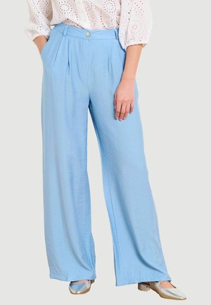 Žena oblečená v svetlomodrých širokých nohaviciach s plisovaním, bielom blúzke s dierkami a metalických strieborných nízkych podpätkoch, stojaca s jednou rukou v vrecku.