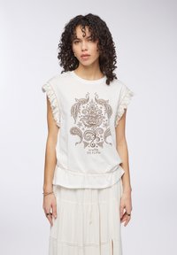 Top bianco senza maniche con dettagli in fringes, caratterizzato da un design a paisley e fiori marrone. Testo "VAI CON IL FLUSSO" al centro.