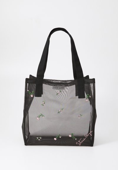 Black mesh tote bag with embroidered floral patterns, featuring sturdy black handles and a logo tag on the front. Translucent material.