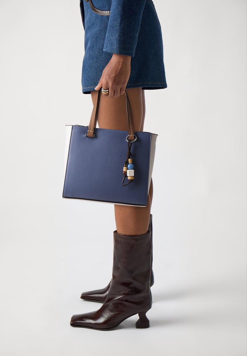 Personne portant des bottes mi-mollet marron foncé et une jupe bleue tenant un sac à main bleu avec des poignées beige et des perles décoratives.