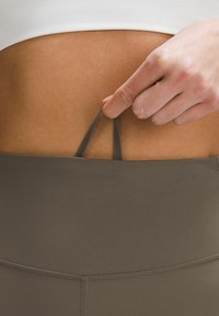 Brązowe legginsy z wysokim stanem z teksturowanej powierzchni, na których widać rękę ciągnącą za regulowany pasek w okolicy pasa.