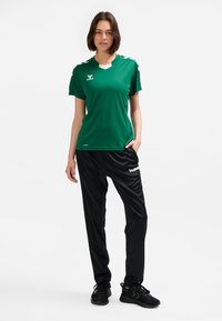 Green athletic jersey with short sleeves and a v-neck, paired with black tapered pants. Both made of breathable fabric; logo on the chest and leg.