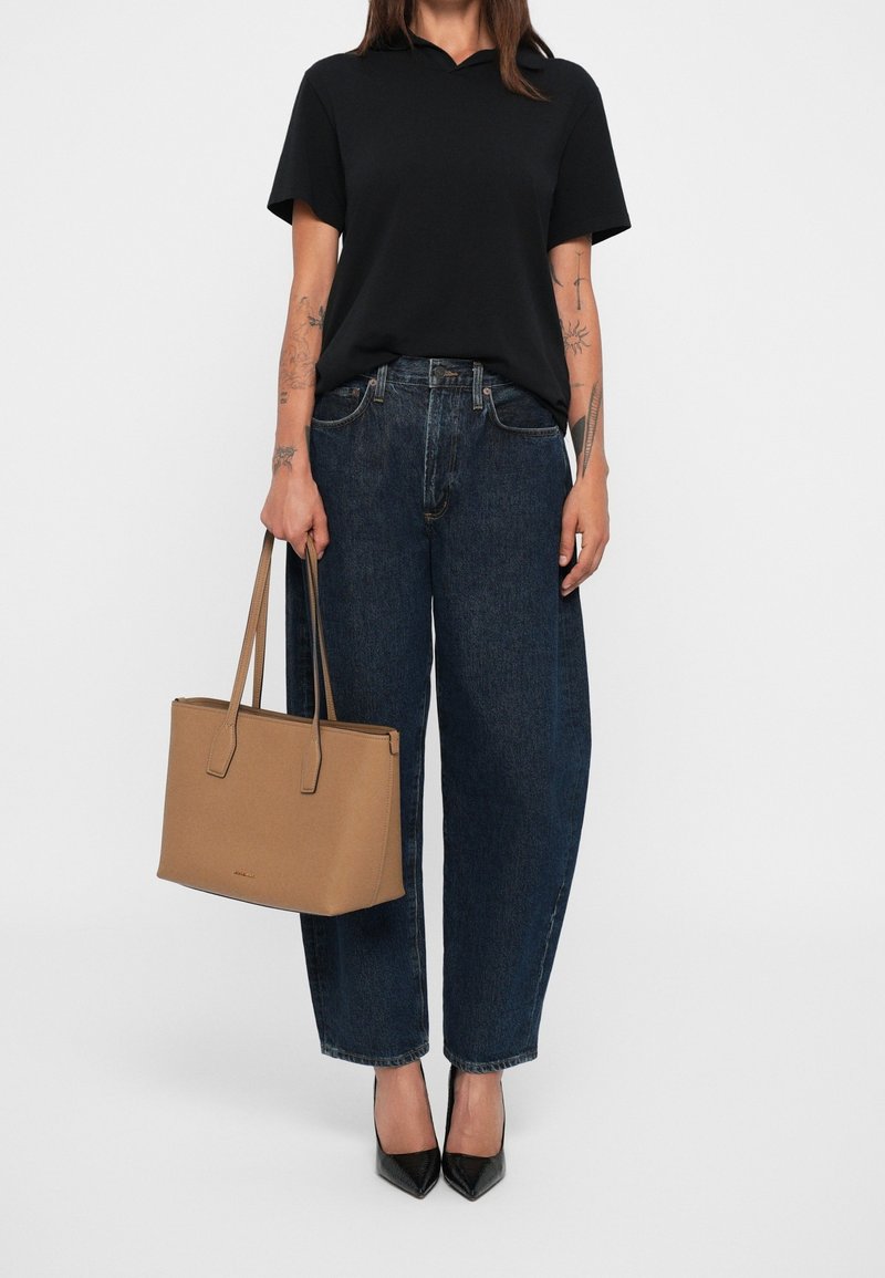 Personne portant une chemise noire à manches courtes, un jean large taille haute bleu foncé, des talons noirs pointus, tenant un sac à main rectangulaire beige.