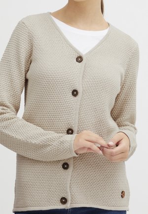 Beige strukturierte Strickjacke, zugeknöpft über einem weißen Hemd, mit langen Ärmeln und einem kleinen runden Aufnäher nahe dem Saum.