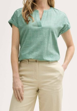 Femme portant un haut vert à manches courtes avec un col en V et un pantalon beige taille haute, une main dans la poche, fond neutre.