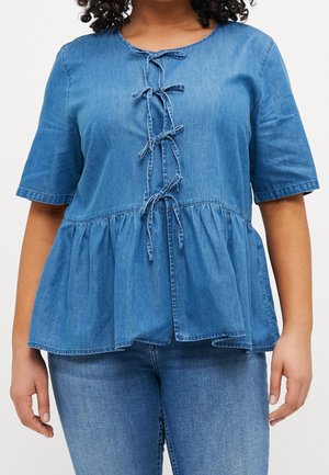 Vrouw draagt een blauwe denim peplum top met korte mouwen en vijf geknoopte strikken aan de voorkant, gecombineerd met een blauwe spijkerbroek.