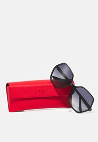 Black hexagonal sunglasses with gradient lenses, placed on a red textured case embossed with the brand "GUESS."