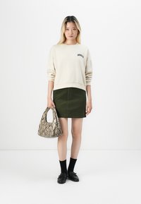 Sweatshirt crème avec le texte "BROOKLYN", jupe en velours côtelé verte, sac à main en imprimé serpent texturé, chaussettes noires et mocassins. Tenue simple et décontractée.