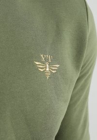 Oliwkowo zielony sweter o miękkiej fakturze, z małym złotym haftowanym logo pszczoły i inicjałami po lewej stronie klatki piersiowej.