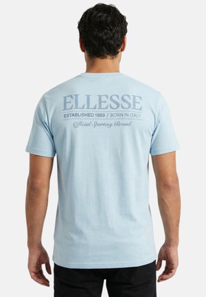 Homme portant un T-shirt bleu clair avec l'inscription "ELLESSE Established 1959 / Born in Italy Official Sporting Brand" dans le dos, face tournée à l'opposé.