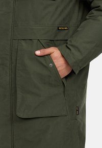 Parka verde oliva con tasca frontale, dettagli cuciti e etichetta del logo. Il tessuto sembra liscio e resistente, adatto per l'uso all'aperto.