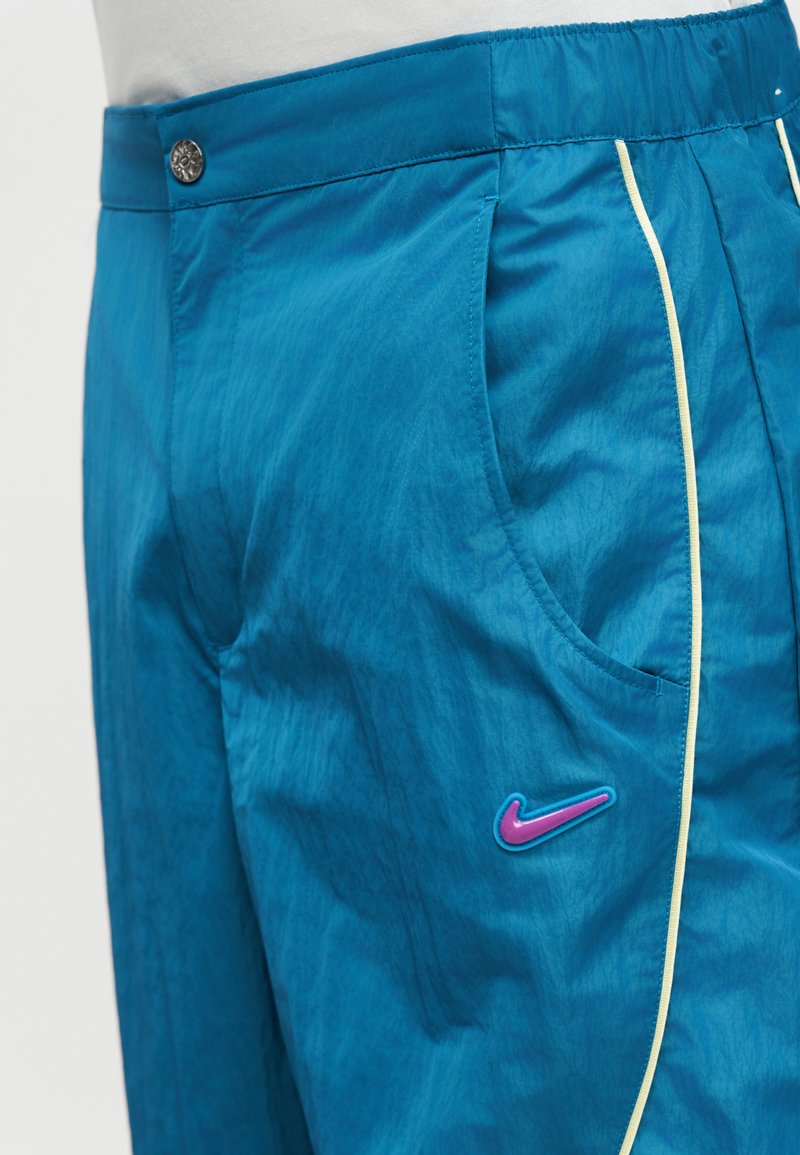 Pantaloni sportivi blu con piping giallo, tasche laterali, chiusura con bottoni e logo Nike Swoosh rosa e blu sulla coscia.