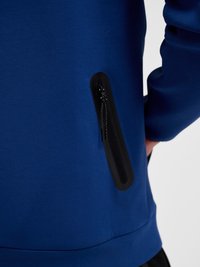 Giacca blu con tasca verticale con cerniera nera sul lato, che mostra parzialmente una mano appoggiata vicino all'anca.