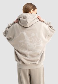 Beżowa, oversize bluza z kapturem z teksturowanym wzorem smoka w białym kolorze na plecach. Miękka tkanina, duży kaptur, prążkowane mankiety i dół.