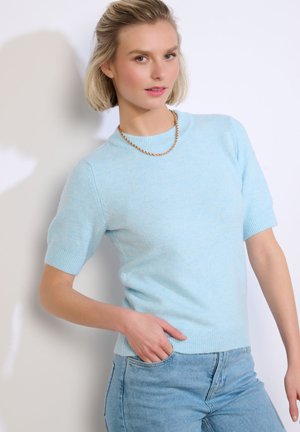 Vrouw met kort blond haar, gekleed in een lichtblauwe trui met korte mouwen, een gouden ketting en lichtblauwe spijkerbroek, poseert tegen een witte achtergrond.