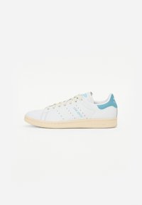 Hvide sneakers med lyseblå accenter, perforerede sidelister og en tekstureret gummisål. Har "STAN SMITH" branding på siden.