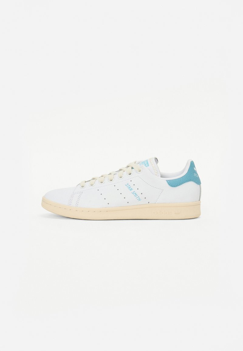 Hvide sneakers med lyseblå accenter, perforerede sidelister og en tekstureret gummisål. Har "STAN SMITH" branding på siden.