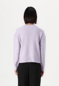 Frau mit kurzen dunklen Haaren, die einen pastellvioletten Pullover und schwarze Hosen trägt, steht vor einem weißen Hintergrund.