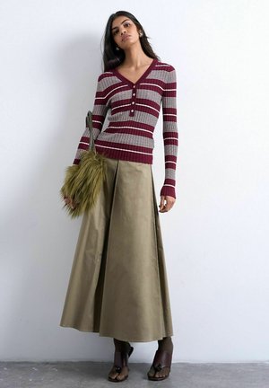 Femme portant un haut à manches longues rouge et blanc rayé, une longue jupe beige, des sandales foncées et tenant un sac à main vert olive en fourrure.