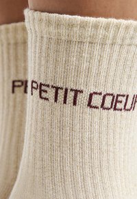 Beige gerippte Socken mit einem subtilen goldenen Faden und weinrotem Schriftzug "PETIT COEUR" am oberen Rand. Weicher Textur.