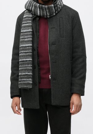 Personne portant une veste en laine gris foncé par-dessus un pull bordeaux, avec une écharpe en tricot rayée grise enroulée autour du cou.