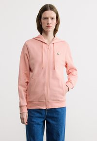 Νεαρή γυναίκα με κοντά καστανά μαλλιά, φορώντας ένα ροζ φούτερ με φερμουάρ και μπλε τζιν, στέκεται μπροστά σε ένα απλό λευκό φόντο.