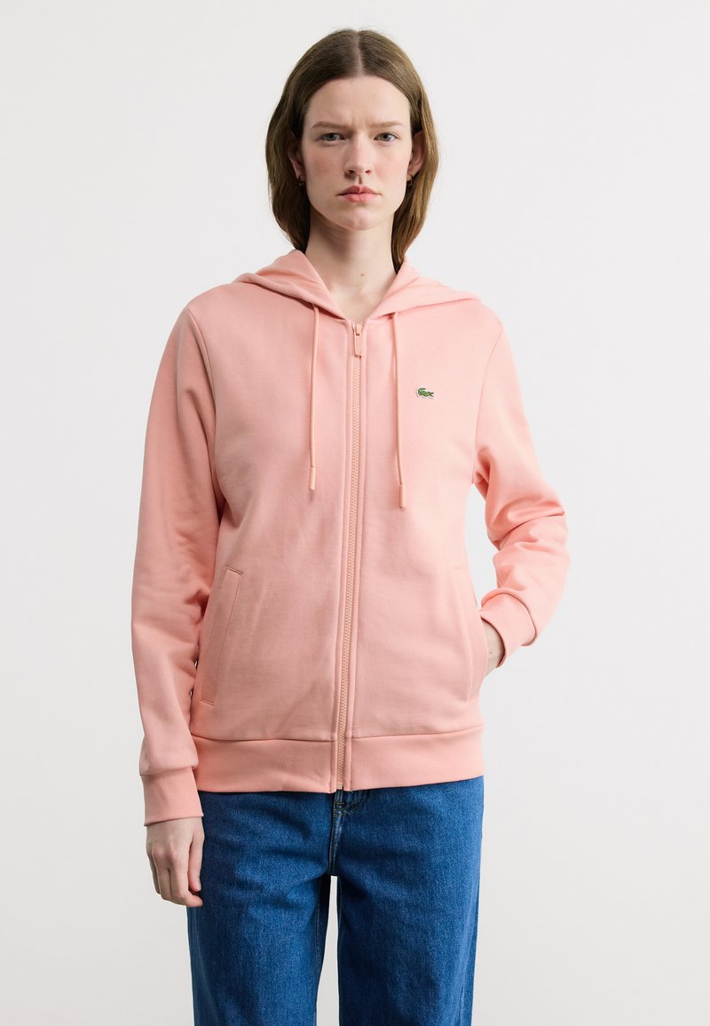 Νεαρή γυναίκα με κοντά καστανά μαλλιά, φορώντας ένα ροζ φούτερ με φερμουάρ και μπλε τζιν, στέκεται μπροστά σε ένα απλό λευκό φόντο.