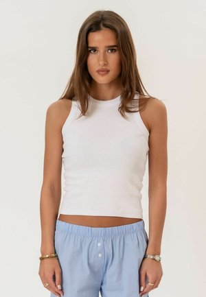 Młoda kobieta o długich brązowych włosach, ubrana w biały top bez rękawów i jasnoniebieskie spodnie, stojąca na jednolitym tle.