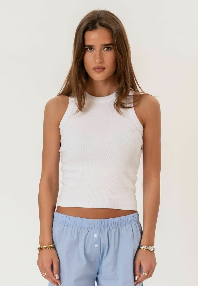 Jeune femme aux longs cheveux bruns portant un haut blanc sans manches et un pantalon bleu clair, debout devant un fond uni.