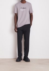 Lysegrå bomulds t-shirt med "Calvin Klein" logo, parret med sorte bukser og sort fodtøj, stående mod en neutral baggrund.