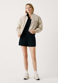 Lichtbeige varsityjack met geborduurd "R" en cijfers, gecombineerd met een zwarte top en shorts, tegen een minimalistische achtergrond.