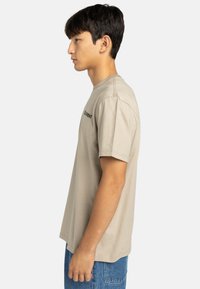 Béžové bavlnené tričko s krátkymi rukávmi a okrúhlym výstrihom, s jemným čiernym logom na ľavej hrudi. Model stojí v profile.