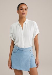 Camicia bianca a maniche corte con textura e colletto abbottonato, abbinata a una gonna avvolgente in denim blu chiaro con dettagli di bottoni laterali.