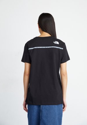 Pitkät tummahiuksinen henkilö pukeutuu mustaan The North Face -t-paitaan, jossa on logo ja merkki yläselässä, yhdistettynä siniseen farkkuhameeseen.