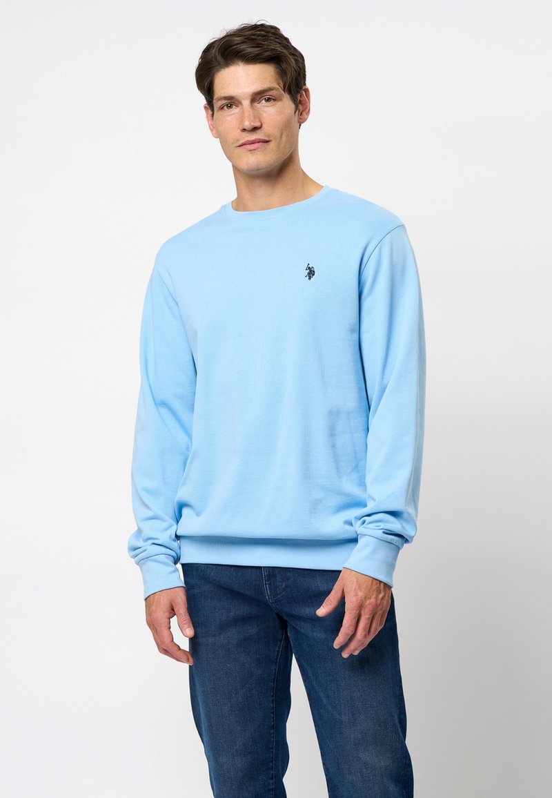 Svetlo modra dolga majica iz mehkega materiala, z okroglo ovratnikom in majhnim črnim logotipom na levem prsnem delu. Kombinirana z džinsami.