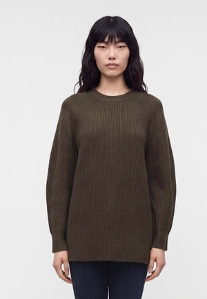 Femme aux longs cheveux noirs portant un pull en maille côtelée olive foncé et un pantalon sombre, debout devant un fond clair uni.