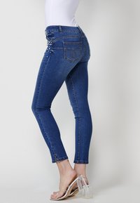 Denim skinny jeans in dark blue with a high waist, featuring pearl embellishments on the back pocket, and ankle-length cut. Clear heeled sandals.