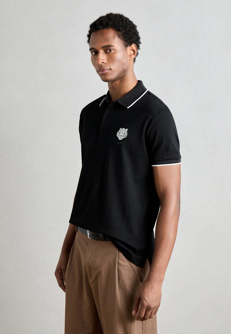 Zwarte poloshirt met witte bies, voorzien van een geborduurd logo op de borst, gemaakt van gladde stof, gecombineerd met lichtbruine broeken.