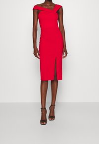 Kobieta w czerwonej, odkrywającej ramiona midi sukience z rozcięciem z przodu i czarnych sandałach na kostkę, stojąca na tle jednolitego białego tła.