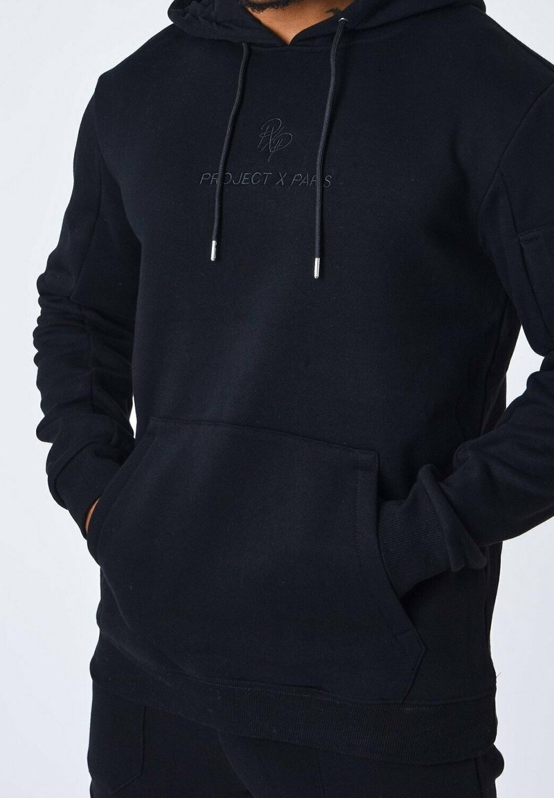Homme portant un sweat à capuche noir avec poche avant et logo "PROJECT X PARIS", mains dans les poches, avec les cordons suspendus.