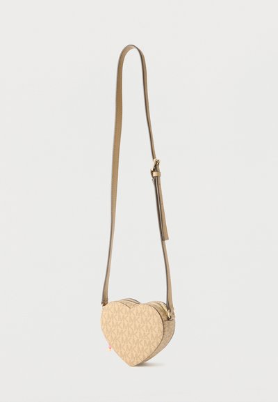 Μπεζ τσάντα χιαστί σε σχήμα καρδιάς με μοτίβο μονόγραμμα και ρυθμιζόμενο λουρί κρεμασμένη σε λευκό φόντο.