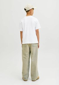 Witte katoenen t-shirt, losvallend; lichtgroene denim broek met achterzakken; beige pet; tan schoeisel; effen achtergrond.