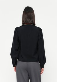 Blusa preta de mangas longas com gola, apresentando mangas bufantes e um tecido texturizado. A vista de trás mostra um caimento relaxado e um acabamento suave.