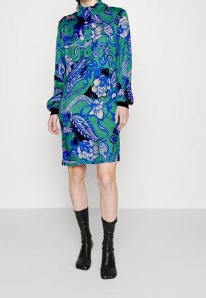 Femme portant une robe à manches longues à motifs floraux verts, associée à des bottes en cuir noires montant jusqu'au milieu du mollet, sur un fond clair uni.