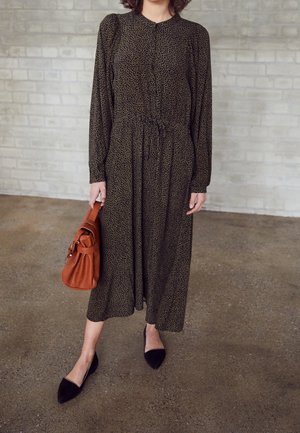 Vestido de manga larga con patrón en negro y dorado, cintura ceñida, combinado con un bolso marrón y zapatos planos negros puntiagudos sobre un suelo texturizado.